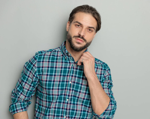 handsome bearded man looking forward while adjusting shirt collar in front of grey background in studio - Photo, Image