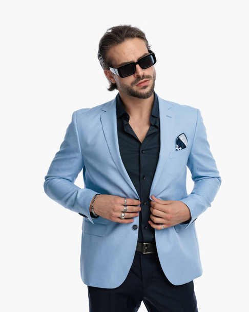 cooler Modetyp mit Sonnenbrille, der wegschaut, während er Anzug vor weißem Hintergrund knöpft  - Foto, Bild