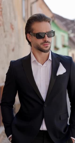 homme d'affaires de mode cool avec des lunettes de soleil portant un costume avec chemise à col ouvert tenant la main dans les poches et posant d'une manière confiante tout en étant à l'extérieur dans une ville médiévale de Roumanie - Séquence, vidéo