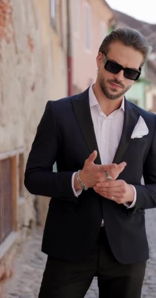 sexy bearded man in suit with untied shirt and sunglasses touching hands and looking to side in front of old city from Romania - Footage, Video