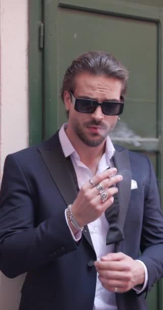 sexy hombre elegante con corbata desatada y gafas de sol mirando a un lado y fumar mientras está fuera en una antigua ciudad medieval - Imágenes, Vídeo