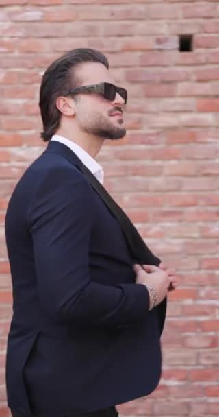 side view of attractive young businessman with sunglasses looking behind while closing suit and walking outdoor in a medieval city from Transylvania, Romania - Footage, Video