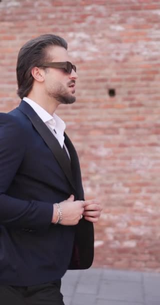 elegant fashion businessman adjusting suit and sunglasses and walking while looking to side and behind and being outdoor in an old city - Footage, Video