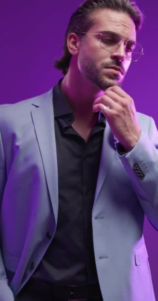 atraktivní módní muž s brýlemi dotýkající se brady, hledající na stranu a upravující oblek před šedým pozadím s barevnými světly - Záběry, video