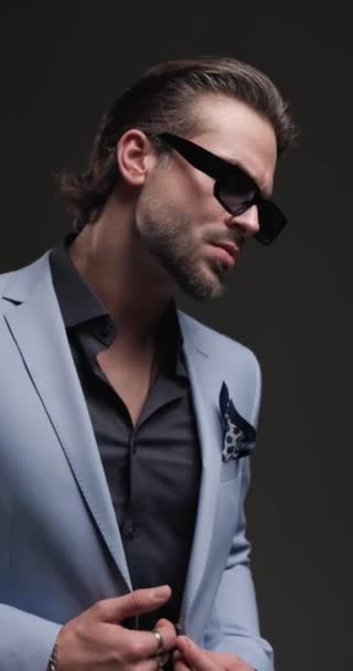 aantrekkelijke elegante zakenman met een zonnebril hand in hand, op zoek naar opzij en wrijven handen op grijze achtergrond met lichten op - Video