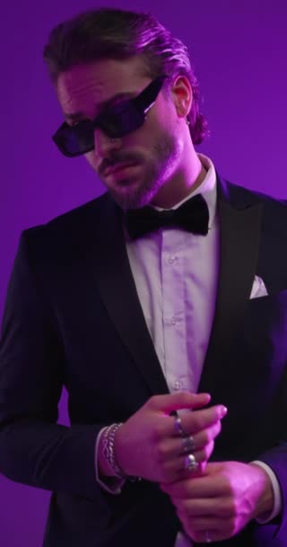 elegant bearded businessman fixing tuxedo, looking to side, touching lips in a seductive way and turning around on colorful lights background - Footage, Video