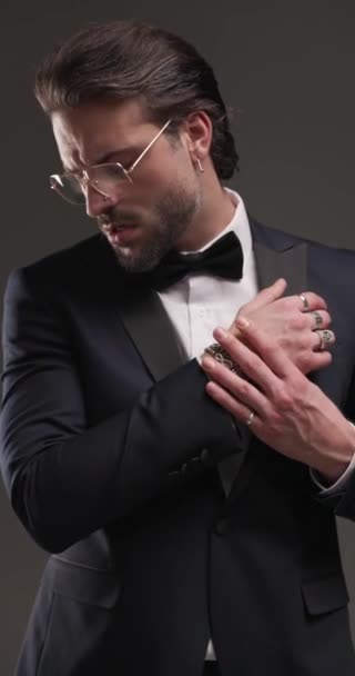 attraktiver eleganter Mann im Smoking, der nach unten schaut, Armbänder und Tux befestigt, nach unten schaut, sich umdreht und das Kinn auf grauem Hintergrund berührt, mit eingeschaltetem Licht - Filmmaterial, Video