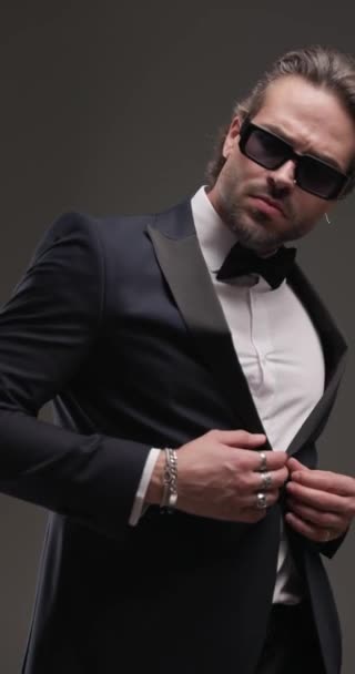 fashion elegant businessman with sunglasses looking to side and adjusting tuxedo, touching lips in a seductive way on grey background - Footage, Video