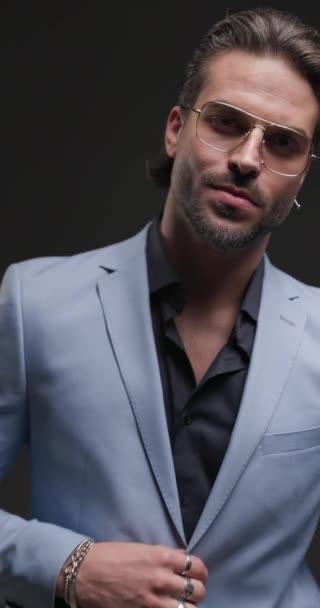 pohledný elegantní muž v obleku s rozhalenou límcovou košilí a brýlemi hledající stranou a odcházející s rukama v kapsách na šedém pozadí - Záběry, video