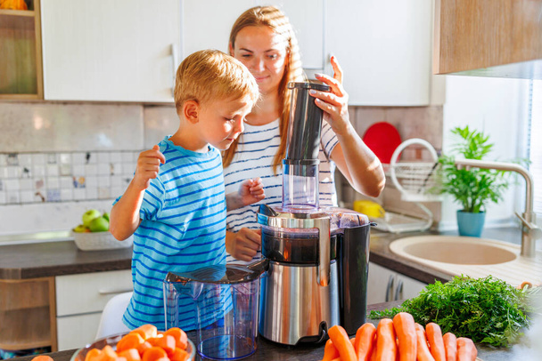 Μικρό παιδί με μητέρα φτιάχνει φρέσκο χυμό καρότου με αποχυμωτή σε μια ηλιόλουστη κουζίνα - Φωτογραφία, εικόνα