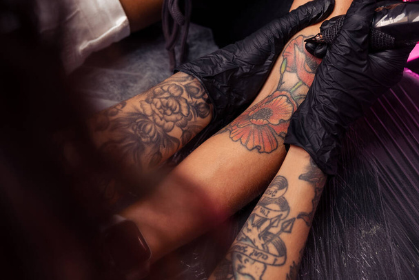 Profesjonalny mistrz tatuażu w czarnych gumowych rękawiczkach tworzący kolorowe arcydzieło artystyczne na kobiecym ramieniu klienta w salonie, kadrowane ujęcie - Zdjęcie, obraz