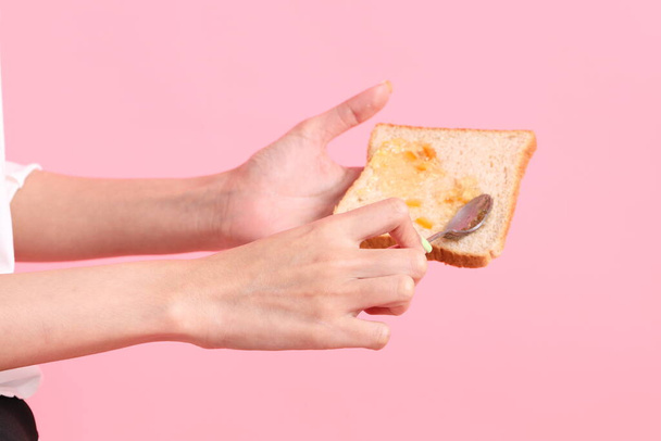 Азиатка наполняет апельсиновый джем нарезанным хлебом на розовом фоне. - Фото, изображение