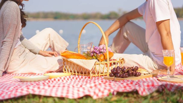 Birbirine aşık genç bir çift kahvaltı sepeti ile battaniyeye oturmuş birlikte piknik yapmak için ekmek ve meyve yiyor.. - Fotoğraf, Görsel