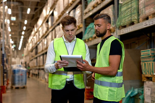 Retail Warehouse full of Shelves with Goods in Boxes, Male Workers Supervisors Holding Paper Tablet Discuss Product Delivery, Dressed in Green Uniform. Distribution Logistics Center - Fotoğraf, Görsel