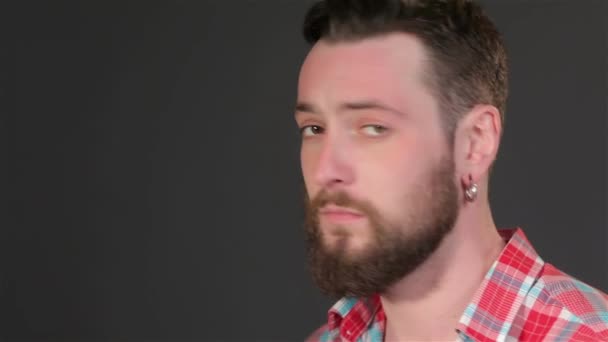 Close up of young hipster making silence gesture. Handsome bearded caucasian man bringing forefinger to his lips. Brunette guy with earrings and fashion hair dress posing isolated on dark gray - Footage, Video