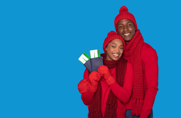 Χειμερινές διακοπές. Ευτυχισμένο νεαρό μαύρο ζευγάρι που κατέχουν διαβατήρια με εισιτήρια κάρτα επιβίβασης, χαμογελώντας στην κάμερα ποζάροντας σε ζεστό κόκκινο κασκόλ και καπέλα σε μπλε φόντο στούντιο, αντίγραφο χώρου - Φωτογραφία, εικόνα