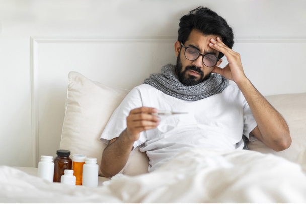 Sick Indian Man Wearing Scarf Sitting In Bed And Holding Thermometer, Young Ill Eastern Guy Measuring Body Temperature, Having Flu Symptoms And Touching Forehead In Frustration, Free Space - 写真・画像