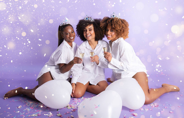 Холостяцкая вечеринка, Праздничный праздник. Три счастливые афроамериканки с коронами и игристыми бокалами, позирующие среди конфетти и воздушных шаров, празднующих Новый год на фиолетовом фоне - Фото, изображение