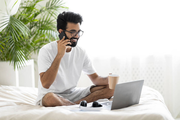 Schöner junger indischer Freelancer Guy, der mit Handy und Laptop spricht, während er im gemütlichen Schlafzimmer auf dem Bett sitzt, der lächelnde Ostler, der mobil telefoniert und Kaffee trinkt und die Remote-Arbeit genießt - Foto, Bild
