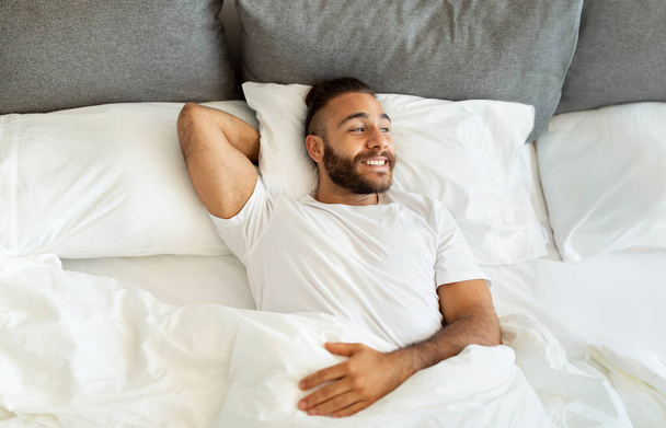 Top view of cheerful attractive bearded young guy hipster wearing white t-shirt chilling in bed in the morning at home. Positive well-rested millennial man lying in bed, look at copy space, smiling - Photo, Image