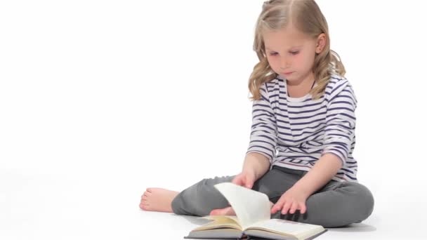 Kaukasisches Kind blättert das Buch isoliert auf weißem Hintergrund. kleines blondes Mädchen mit einem Buch auf dem Boden sitzend. Kleines Kind bewegt die Seiten mit den Händen - Filmmaterial, Video