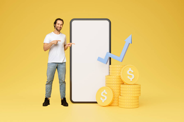 Ευτυχισμένος έμπορος νεαρός άνδρας συνιστώντας την εμπορία mobile app, στέκεται δίπλα σε μεγάλο smartphone με λευκή άδεια οθόνη, mockup, τα διαγράμματα μετοχών και τα κέρματα με το σύμβολο του δολαρίου, δείχνοντας στο τηλέφωνο, κολάζ - Φωτογραφία, εικόνα