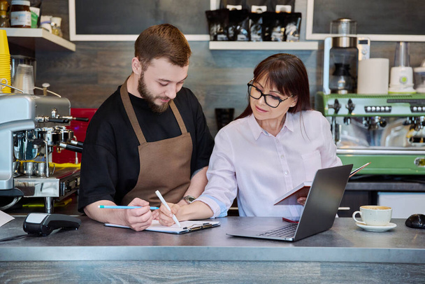 Team, partners, young male and mature woman talking working using laptop standing behind bar in coffee shop. Team, small business, work, staff, cafe cafeteria restaurant, entrepreneurship concept - Photo, image