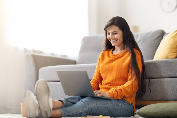 Joyeux attrayant jeune femme hindoue aux cheveux longs freelance travaillant sur ordinateur portable de la maison, assis sur le sol dans le salon. Femme indienne développeur dactylographier sur clavier portable et souriant, espace de copie - Photo, image