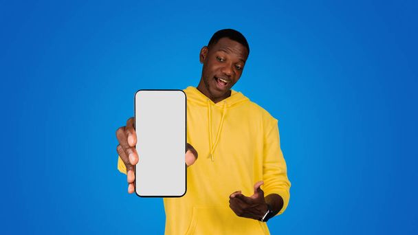 Uśmiechnięty zaskoczony młody czarny mężczyzna wskazując palcem na telefon z pustym ekranem, poleca aplikację, izolowane na niebieskim tle, studio, panorama. Dobre wieści, strona internetowa, reklama i oferta i gadżet - Zdjęcie, obraz