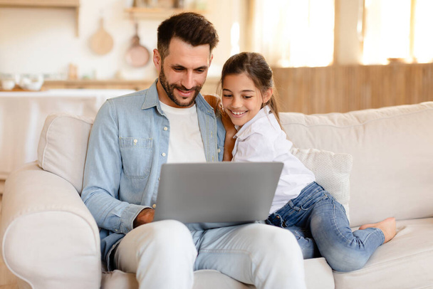 Alegre papá árabe y la pequeña hija se divierten con el ordenador portátil en casa, viendo dibujos animados y videos juntos en línea, relajarse con la computadora gadget en el sofá en la sala de estar moderna - Foto, Imagen
