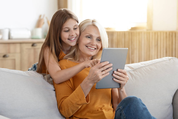 Pozitív örömteli vonzó szőke idős nő 60-as évek szórakozás unokája csinos tini idős lány otthon, ülni a kanapén a nappaliban, a digitális tabletta, játszani mobil játék, szép app - Fotó, kép