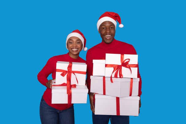 Vánoční donáška. Radostný černošky mladý pár drží mnoho vánoční dárky boxy, Pózování v Santa klobouky a usmívání se na kameru na modrém pozadí, Studio Shot. Zimní dovolená, Nový rok Nabídky - Fotografie, Obrázek