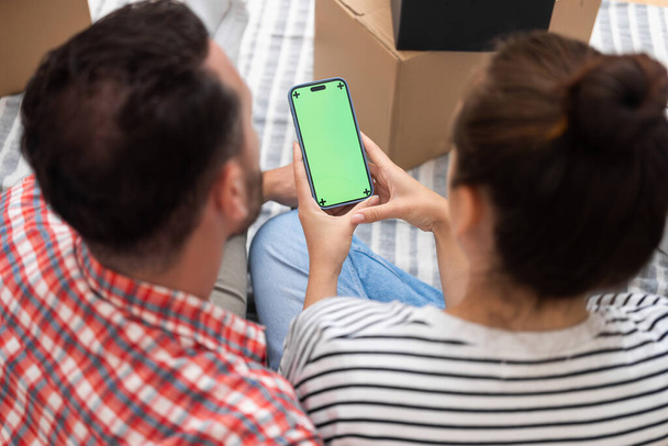 A kezek egy okostelefont tartanak egy zöld modell képernyővel a kartondobozok között, miközben egy fiatal pár áthelyezi magát az új otthonukba. - Fotó, kép