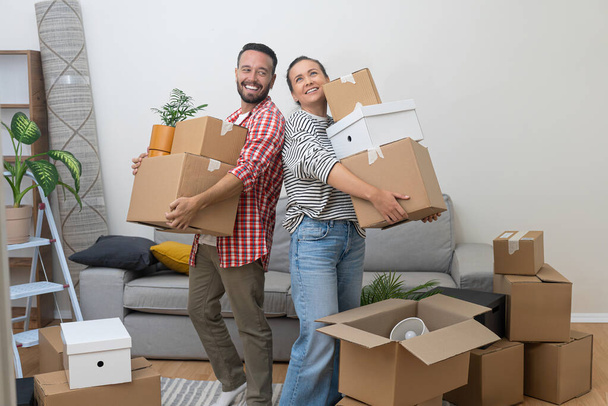 Relocation Challenges: A young couple, homeowners, struggles to hold a pile of cardboard moving boxes as they enter their new living room, determined to make it their own. - Photo, Image