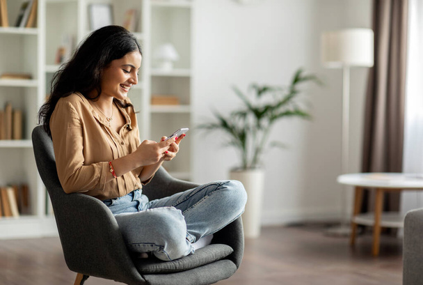 Szczęśliwa Hinduska kobieta korzystająca z telefonu komórkowego, siedząca w fotelu, przeglądająca internet lub pisząca z przyjaciółmi, korzystająca z komunikacji mobilnej w domu, wolnej przestrzeni - Zdjęcie, obraz