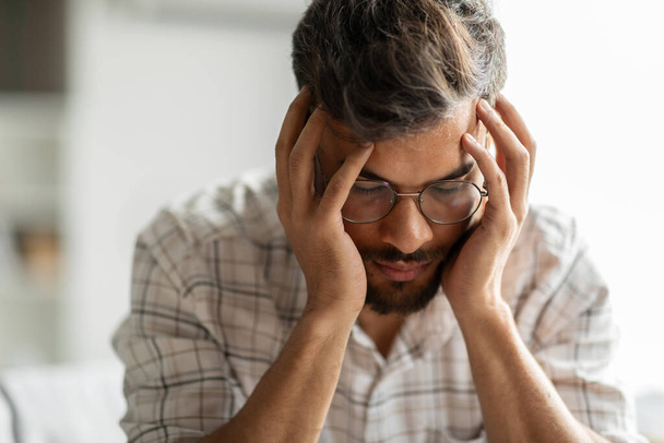 Young indian man suffering from migraine, feeling unwell, touching head with closed eyes, suffering acute headache while sitting at home, copy space - Photo, Image