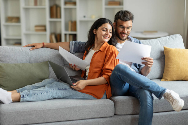 Glückliches indisches Paar prüft Dokumente, Ehepartner lesen Versicherungsvertrag oder Immobilienschein, Mann und Frau sitzen auf Couch im Wohnzimmer, Kopierraum - Foto, Bild