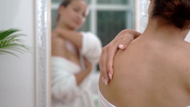 Donna sorridente diffusione lozione idratante sulla pelle della spalla in bagno signora in accappatoio utilizzando prodotto cosmetico per la cura del corpo a casa procedure di trattamento spa - Filmati, video