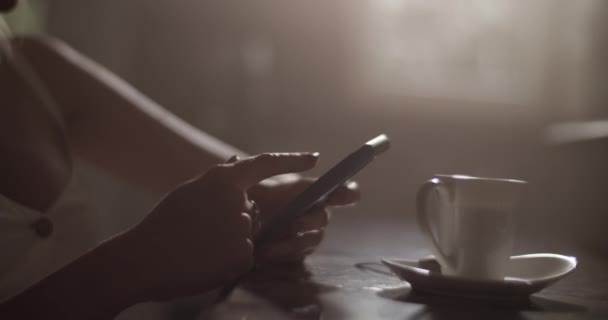 Real time handheld shot of crop anonymous lady sitting at table and using mobile phone while surfing social media during coffee break at home in kitchen with dim light - Footage, Video