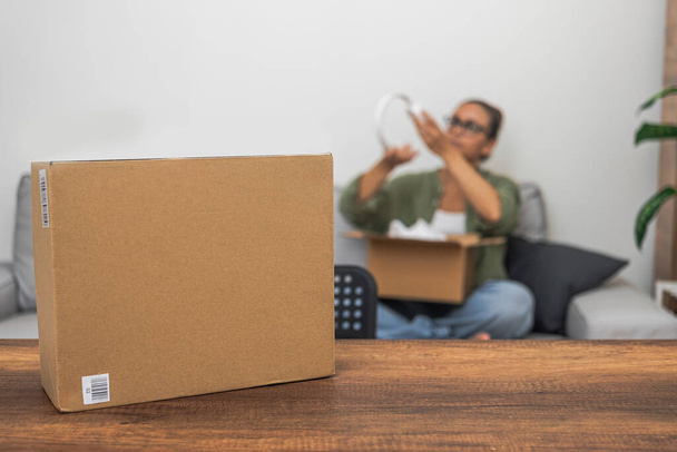 Az asztalon csomagküldő dobozok várnak, míg a háttérben egy nő kibontja az online vásárlási csomagjait, szimbolizálva a szállítási és piaci vásárlás birodalmát. - Fotó, kép