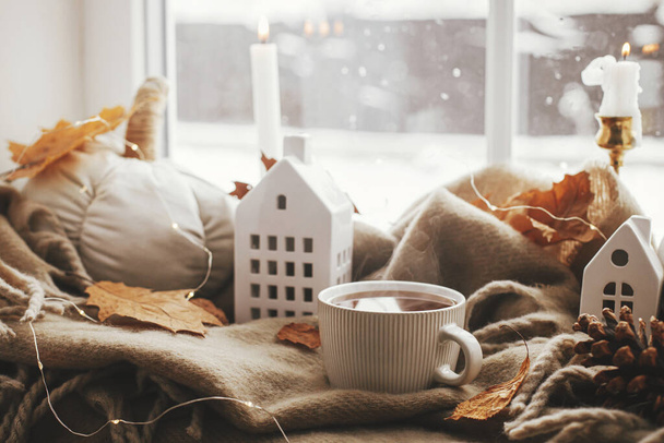Cozy Autumn. Warm cup of tea, stylish pumpkin pillows, fall leaves, candle, lights and cute buildings decoration on brown scarf on windowsill. Autumn hygge, fall home decor - Foto, afbeelding