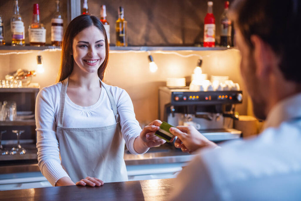 Красивая бариста берет кредитку и улыбается во время работы в баре в кафе
 - Фото, изображение
