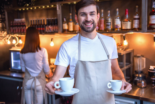 Handsome barista in apron is holding cups of coffee and smiling while working at the cafe - Photo, Image