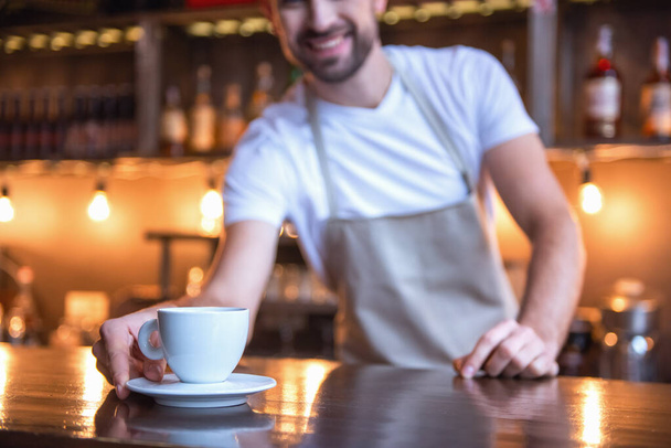 Imagen recortada de barista guapo en delantal sosteniendo una taza de café y sonriendo mientras trabajaba en el mostrador del bar en la cafetería. Copa en foco
 - Foto, Imagen