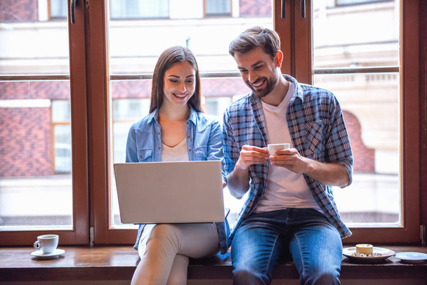 Onnellinen nuori pari käyttää kannettavaa tietokonetta, juo kahvia ja hymyilee istuessaan kahvilassa.
 - Valokuva, kuva