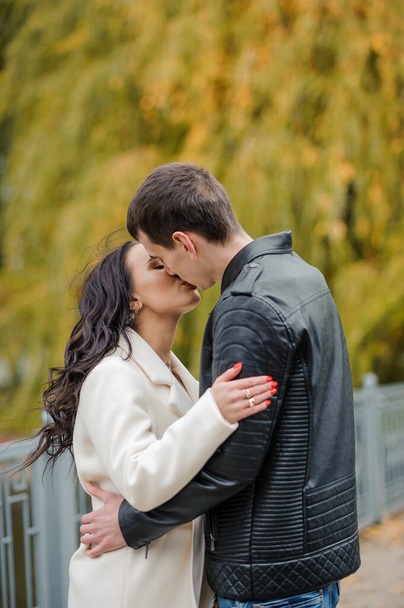Um homem abraça a mulher grávida. Um homem de casaco preto abraça e beija uma mulher de casaco branco. Outono - Foto, Imagem