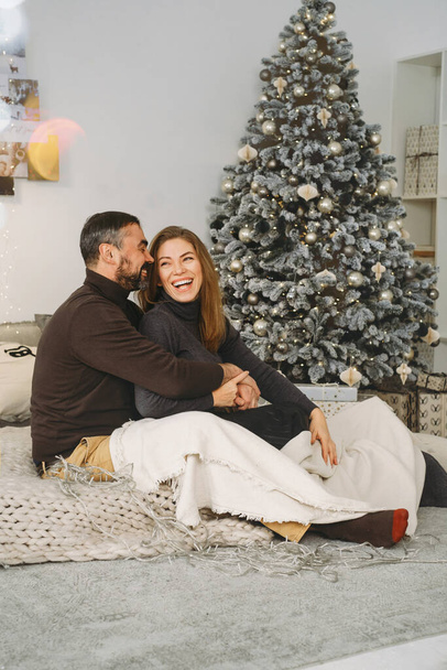 Happy couple celebrating romantic Christmas eve at home. Christmas interior decoration for family party. Copy space, greeting card. Middle-aged man and woman sitting on cozy bed with knitted blanket - Photo, Image