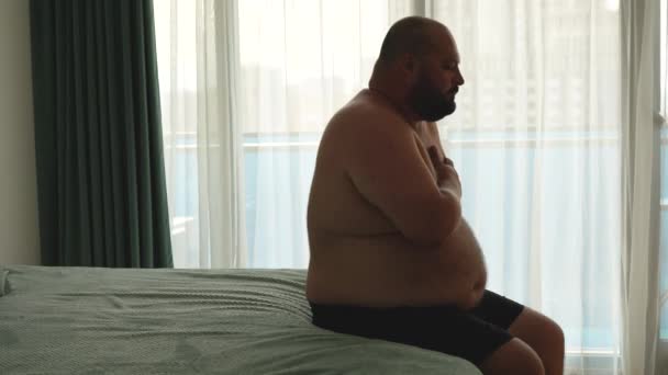 Tlustý muž středního věku cvičí na dýchání, aby se vyrovnal s letní netečností. Nadváha obézní baculatý mladý muž sedící na posteli, těžké dýchání zhluboka zvládnout s hrozným teplem, držení hrudníku - Záběry, video