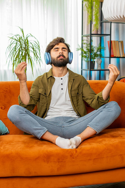 Cálmate, relájate. Hombre árabe de Oriente Medio escuchando música respira profundamente, los ojos cerrados meditando con pensamientos concentrados, mente pacífica. El barbudo se sienta en casa en la habitación en el sofá. Vertical - Foto, imagen