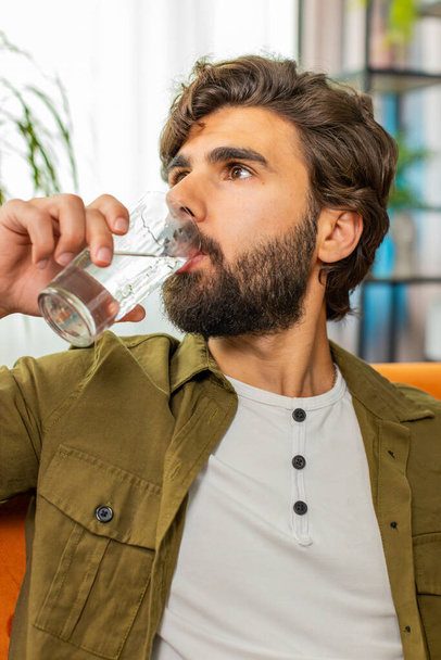 Thirsty man hold glass of natural filtered aqua make sips drinking still water preventing dehydration at home room. Water after reverse osmosis filter. Guy with good life habits, healthy, weight loss - Photo, Image